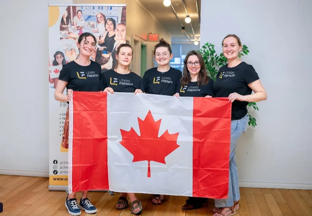 Five women in black Learn French t-shirts stand smiling in an indoor space with wood floors at Learn French Canada